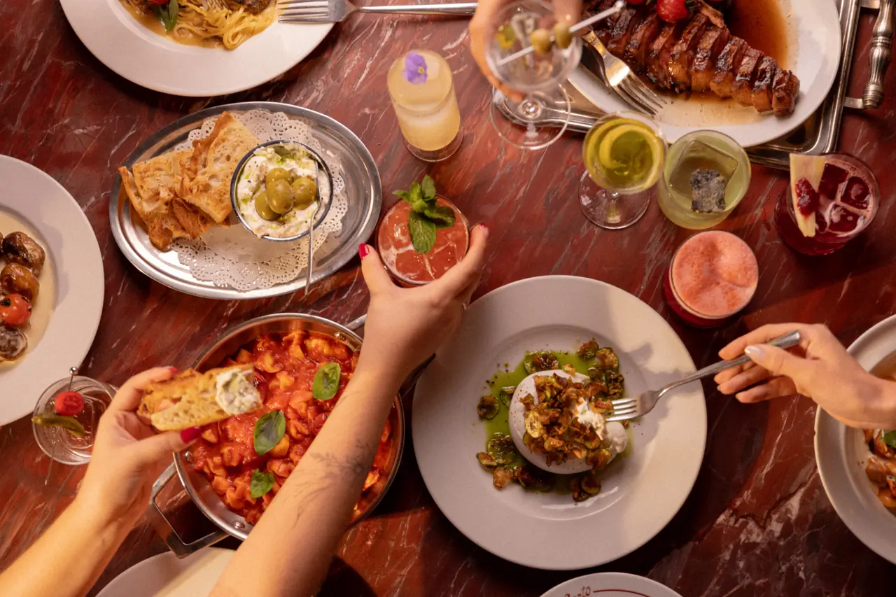 Mesa vista de cima com antepastos, massas, carnes e drinks sendo divididos entre amigos