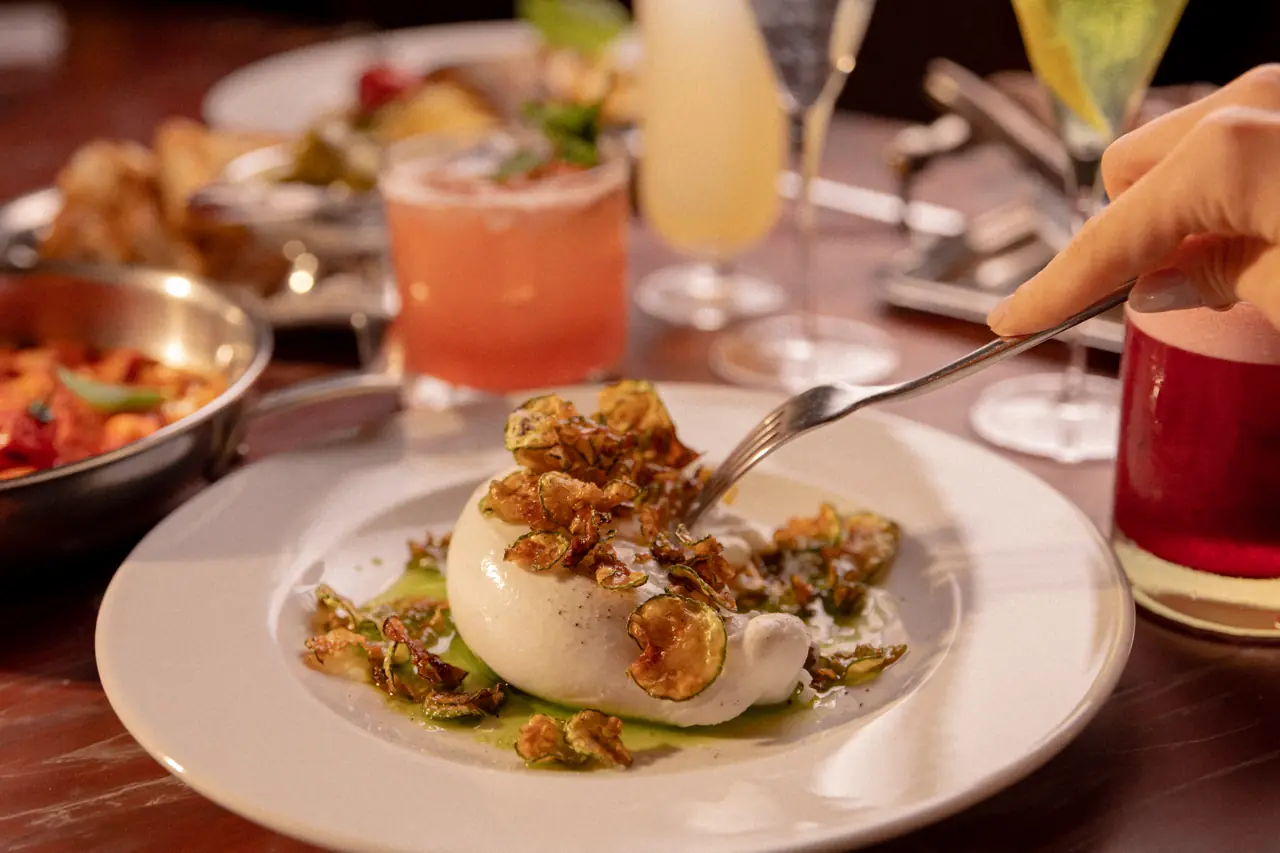 Mãos servindo burrata e antepastos numa mesa de mármore vermelho