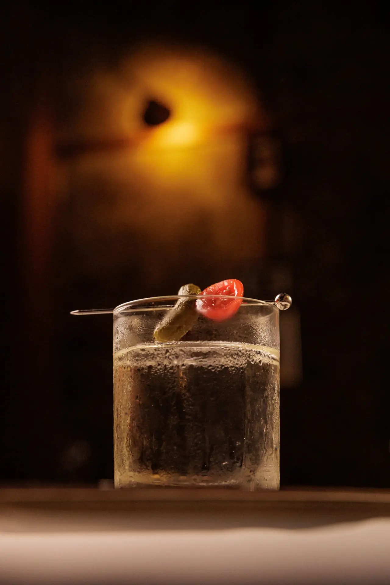 Drink transparente com picles e tomate no palito, contra a luz âmbar do salão