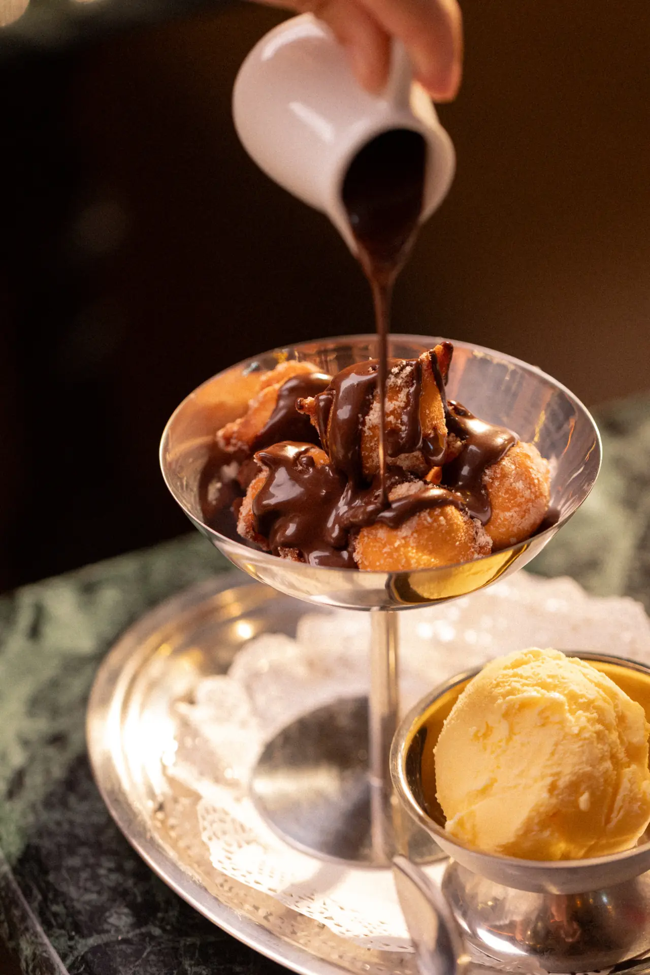 Calda de chocolate sendo despejada sobre profiteroles em taça de prata